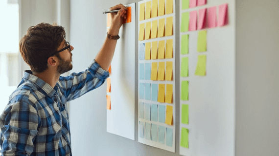 man stands near a large board full of sticky notes, writing on one